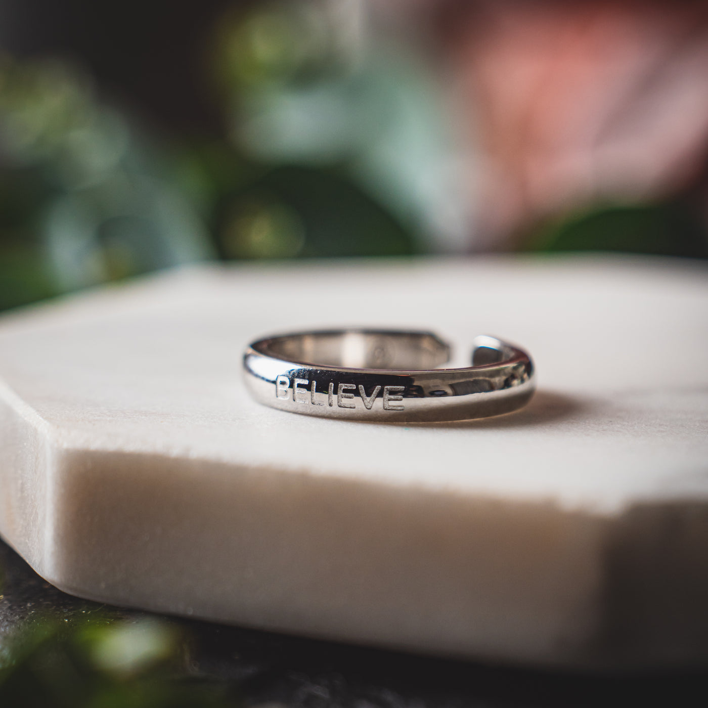 Shiny stainless steel ring with the word 'BELIEVE' engraved, placed on a soft-textured white surface with a blurred background of greenery.