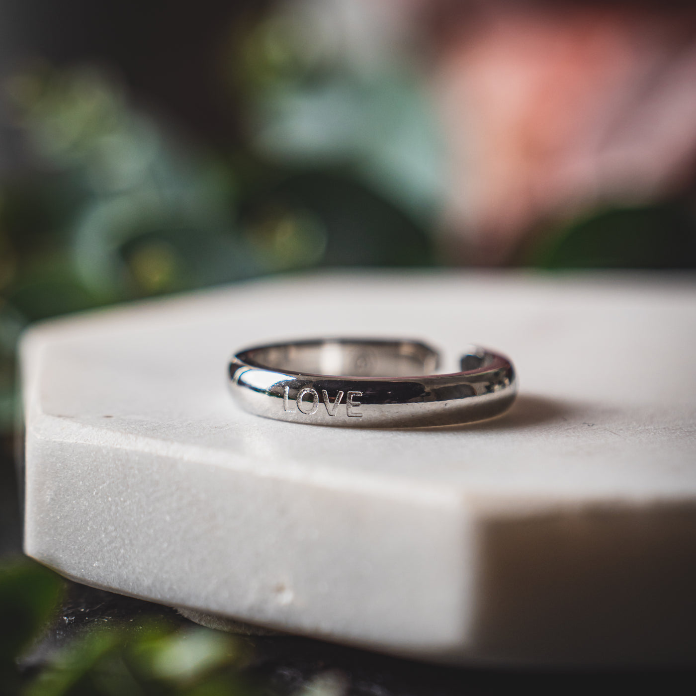 Stainless steel ring with the word 'LOVE' engraved, displayed on a white surface with a blurred green background