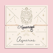 Adjustable peach cord bracelet with a silver 'Aquarius' charm and dangling heart pendant, displayed on a zodiac-themed card with a water bearer illustration and traits reading "Independent, Curious, Bold".