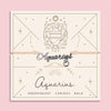 Adjustable peach cord bracelet with a silver 'Aquarius' charm and dangling heart pendant, displayed on a zodiac-themed card with a water bearer illustration and traits reading 