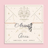 Peach cord bracelet featuring a silver 'Aries' script charm with heart pendant, presented on a zodiac card with ram illustration and personality traits 