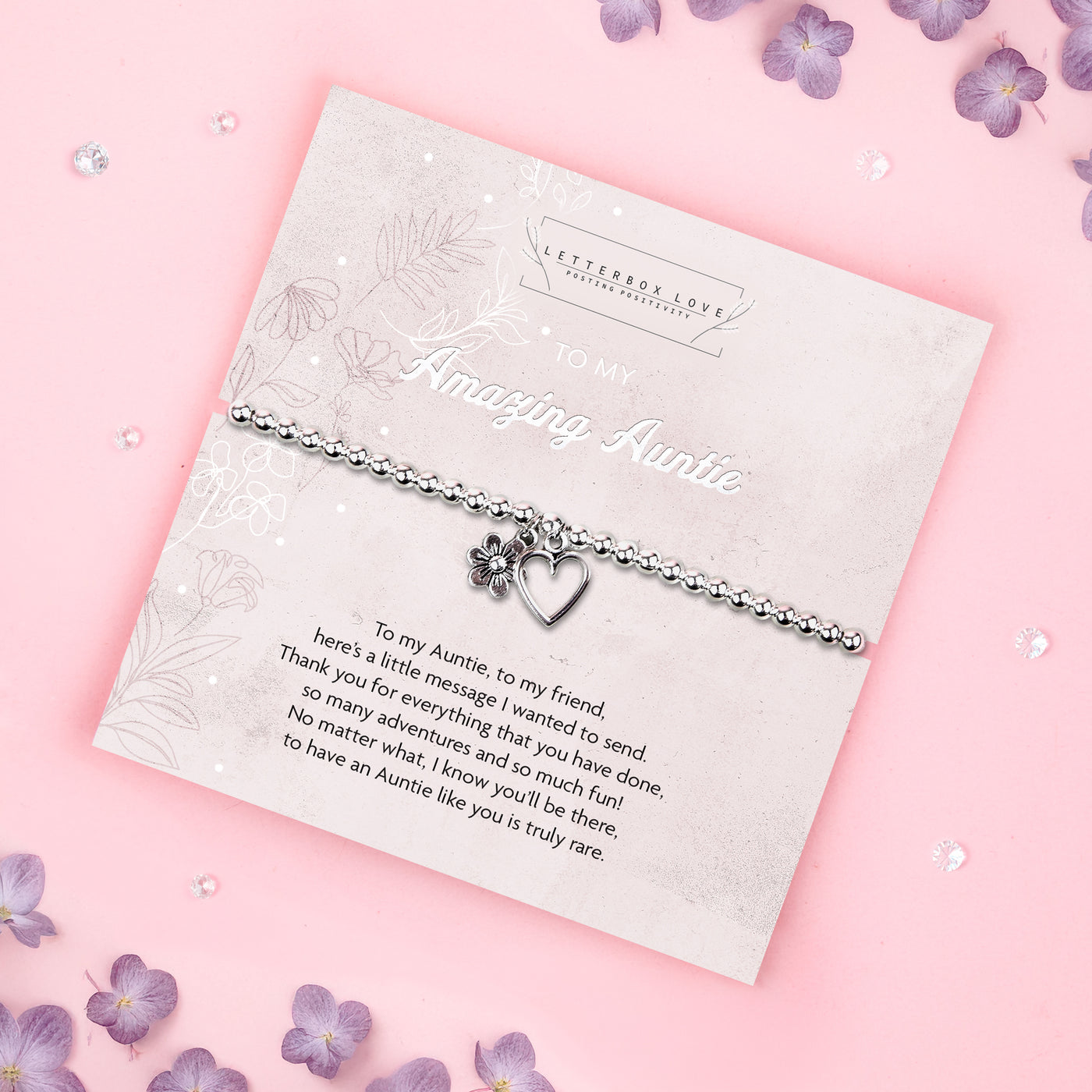 A silver beaded bracelet with a heart and flower charm displayed on a card reading 