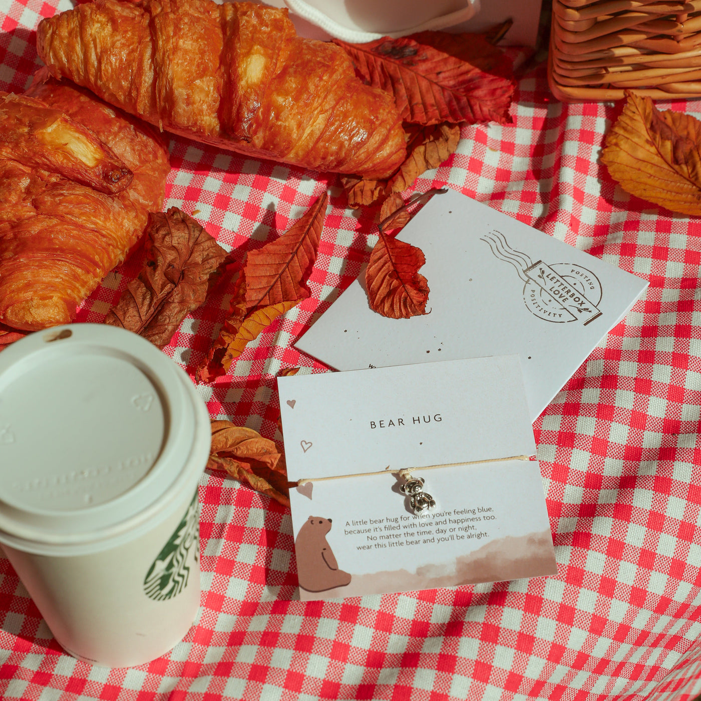 A styled autumn picnic scene with golden croissants on a red and white checkered blanket, scattered dried leaves, a white coffee cup, cards, a wicker basket, and a plush bear toy.