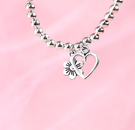 A pale pink background with a silver-plated beaded bracelet dangling with a hollow heart and flower charm. A beautiful gift for someone special.