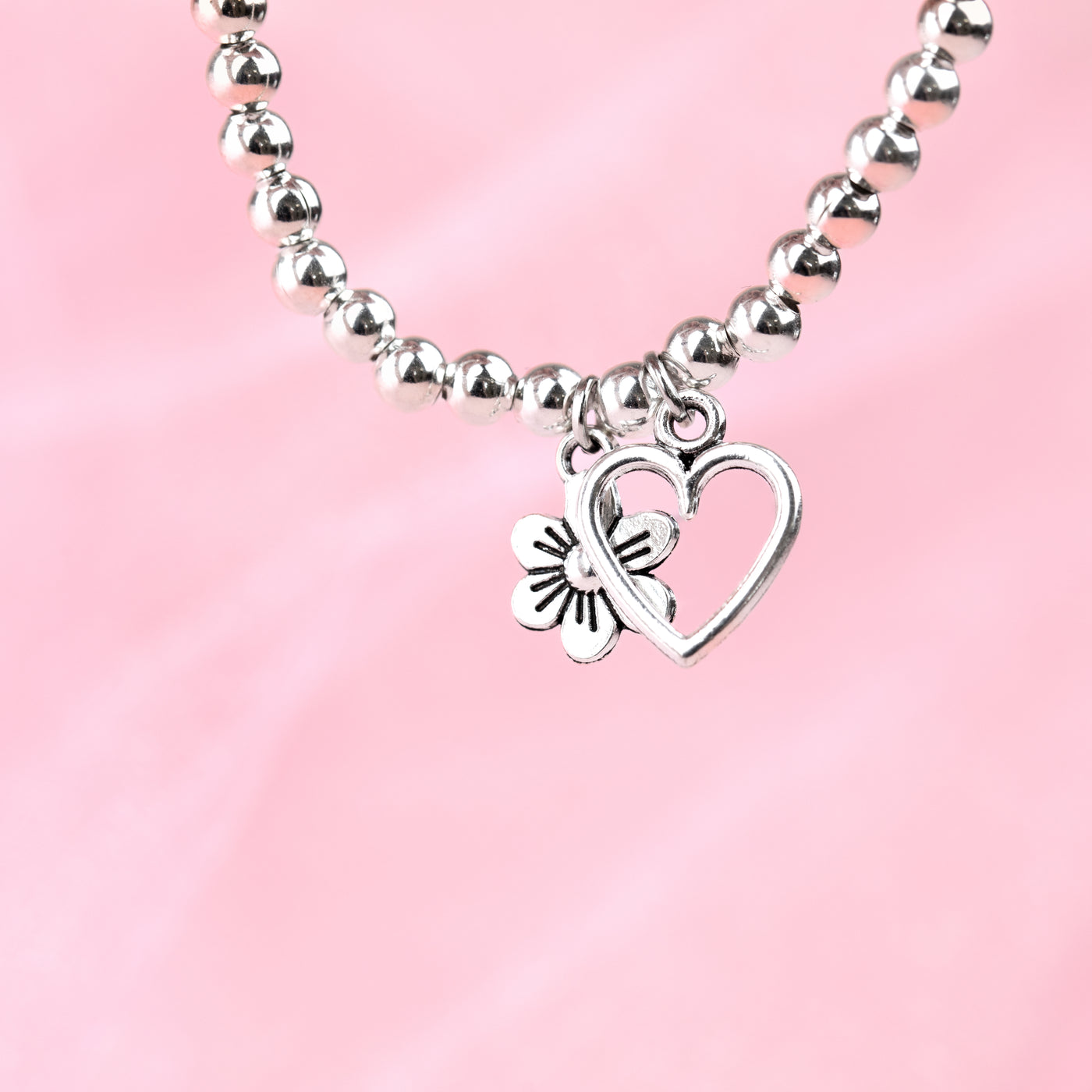 A pale pink background with a silver-plated beaded bracelet dangling with a hollow heart and flower charm. A beautiful gift for someone special.