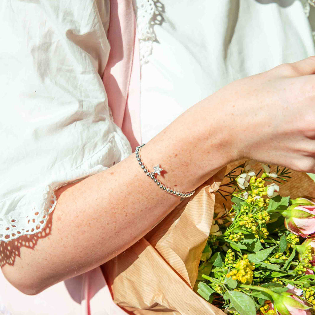 Person holding a bouquet of flowers with a blurred background and a beaded silver bracelet with a beautiful star charm.