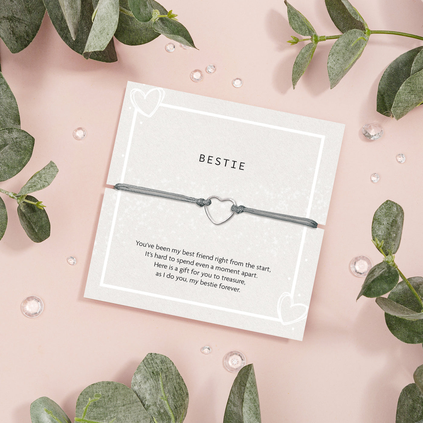 Heart-shaped bracelet on a card with 'BESTIE' text, surrounded by green leaves on a pink background. A sentimental gift for a best friend. 