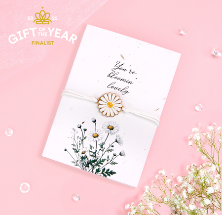 A delicate daisy bracelet featuring a white and yellow flower charm, tied with a soft white cord, displayed on a seeded card with the text 