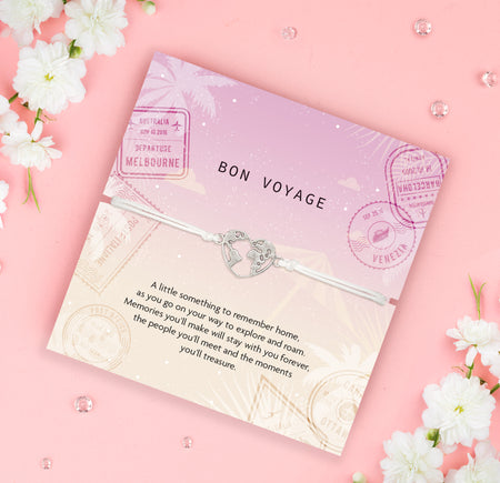 Bon Voyage card with a globe and heart charm on a pink background with white flowers. An affordable travel gift for her