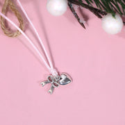 A silver-plated bow and heart charm on a thin pink cord bracelet, displayed on a soft pink background with decorative twine, white berries, and greenery. A way to say thank you at Christmas time.