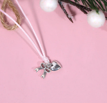 A silver-plated bow and heart charm on a thin pink cord bracelet, displayed on a soft pink background with decorative twine, white berries, and greenery. A way to say thank you at Christmas time.