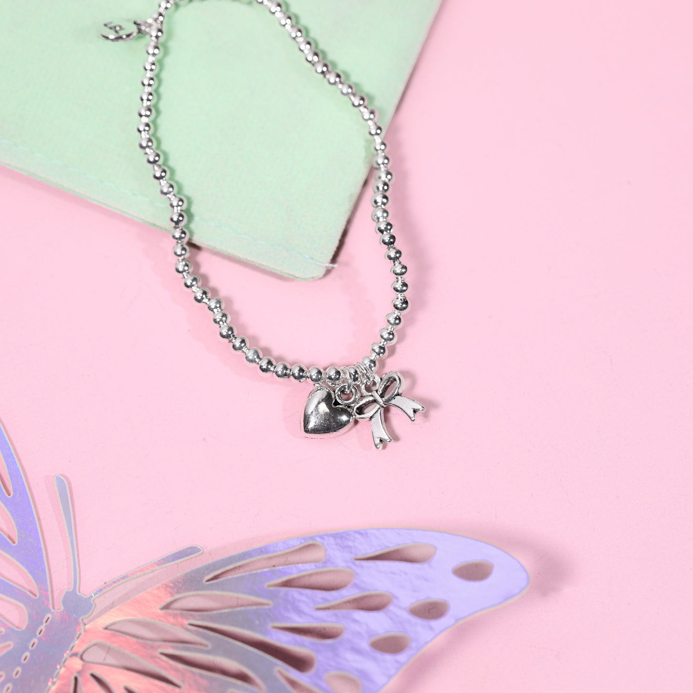 A silver beaded bracelet with a bow and heart pendant, styled on a pastel pink background with a mint green card and a decorative butterfly cutout.