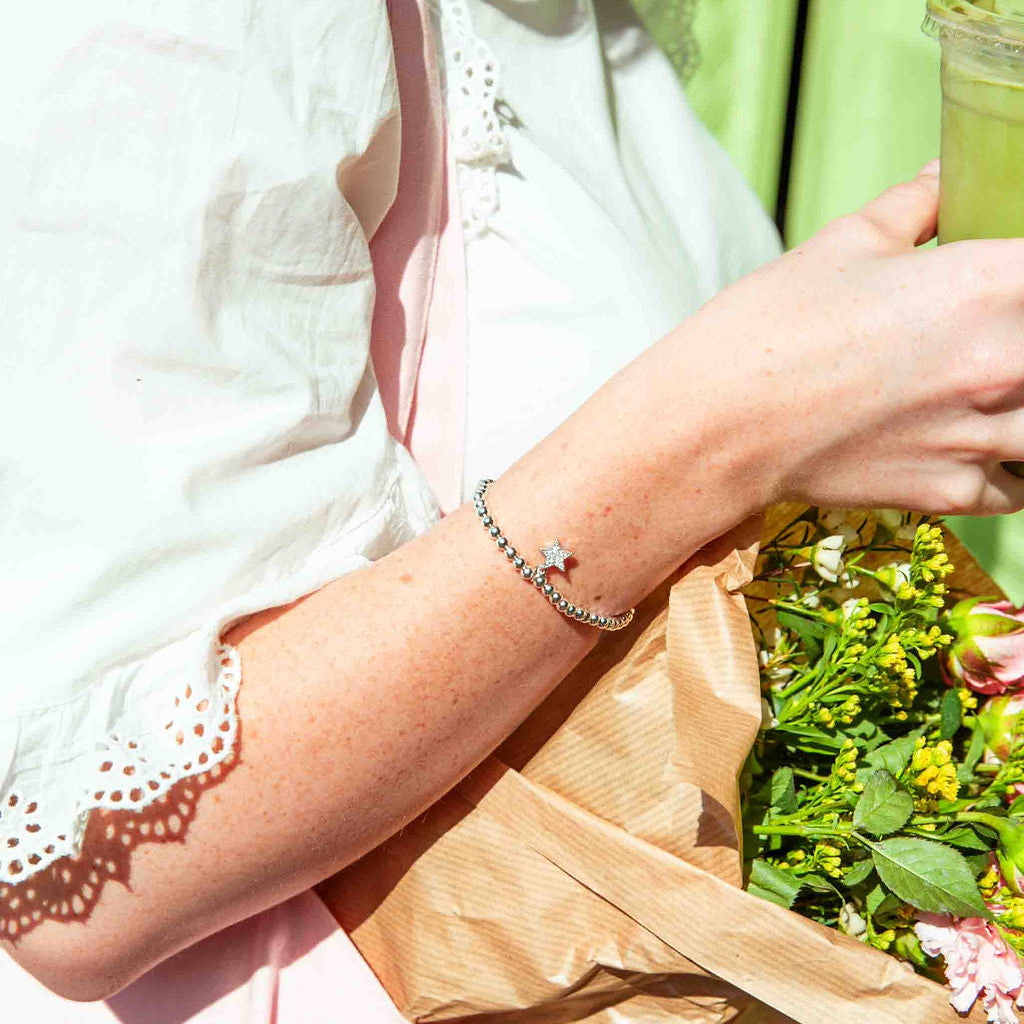 Person holding a matcha and a bouquet of flowers against a green background, she is wearing a silver plated beaded star bracelet
