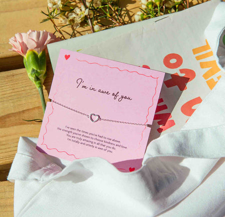 Card with 'I'm in awe of you' message and heart charm on a pink card with red text, surrounded by flowers on a wooden surface.