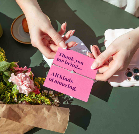 Two hands holding a pink card with a message on a green table with flowers.