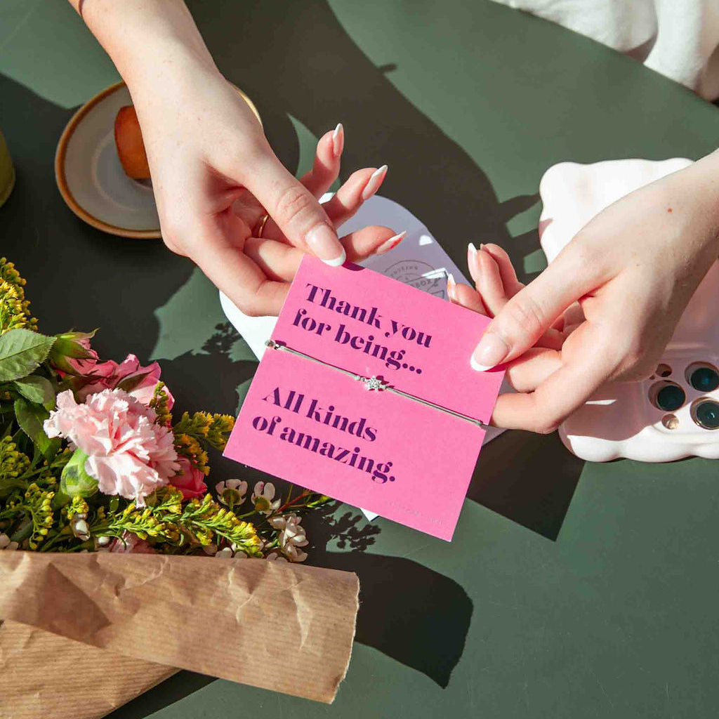 Two hands holding a pink card with a message on a green table with flowers.
