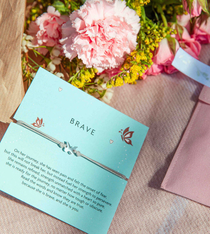 Bracelet on a card with 'BRAVE' text, surrounded by flowers and a pink velvet envelope.