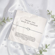 A 'THANK YOU MAID OF HONOUR' appreciation card from Letterbox Love, placed on a silky white fabric background. The card displays a heartfelt message of gratitude and features a delicate silver bracelet with a heart-shaped charm. Set on a silk background.