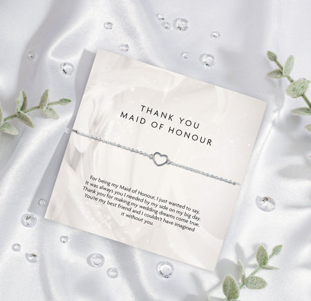 A 'THANK YOU MAID OF HONOUR' appreciation card from Letterbox Love, placed on a silky white fabric background. The card displays a heartfelt message of gratitude and features a delicate silver bracelet with a heart-shaped charm. Set on a silk background.