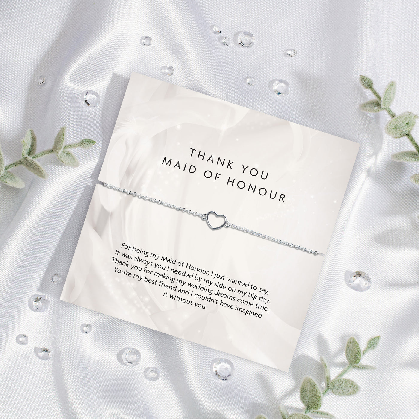 A 'THANK YOU MAID OF HONOUR' appreciation card from Letterbox Love, placed on a silky white fabric background. The card displays a heartfelt message of gratitude and features a delicate silver bracelet with a heart-shaped charm. Set on a silk background.