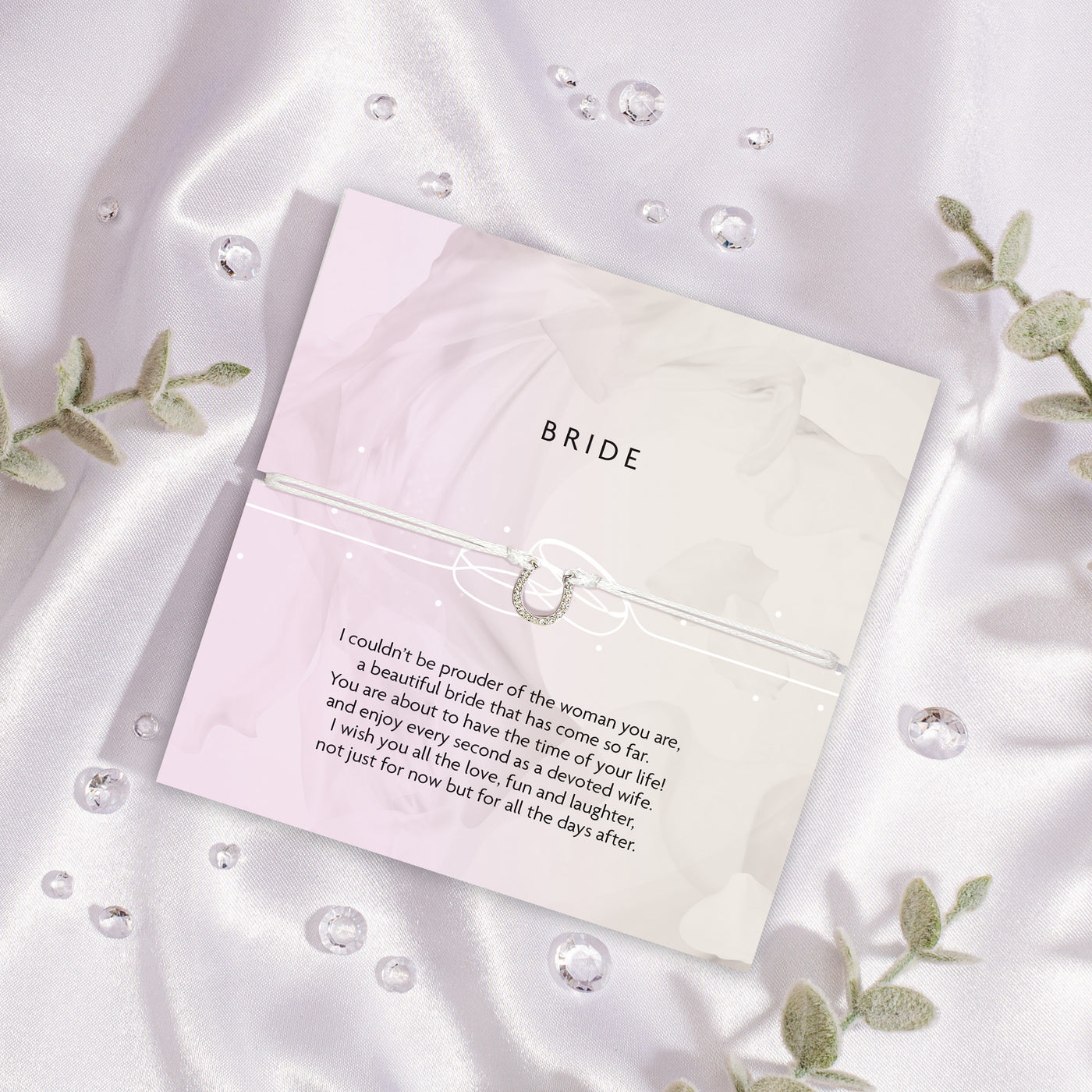 Card for the bride on a satin surface with decorative elements. The bracelet is a white string horseshoe charm. A gift to wish a bride luck in married life.