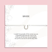 "'BRIDE' silver horseshoe-shaped pendant bracelet on a delicate cord, displayed on a white card with subtle floral designs. The card carries a heartfelt message expressing pride and well wishes for a bride, emphasizing love, fun, and the future journey as a wife."