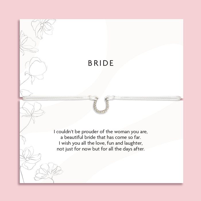 "'BRIDE' silver horseshoe-shaped pendant bracelet on a delicate cord, displayed on a white card with subtle floral designs. The card carries a heartfelt message expressing pride and well wishes for a bride, emphasizing love, fun, and the future journey as a wife."