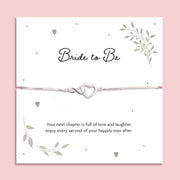 Silver zirconia heart and infinity charm bracelet displayed on a white card with the words "Bride to Be" and a romantic poem, symbolising love and celebration for an upcoming wedding. The bracelet is dainty and adjustable, styled on a soft pink background.