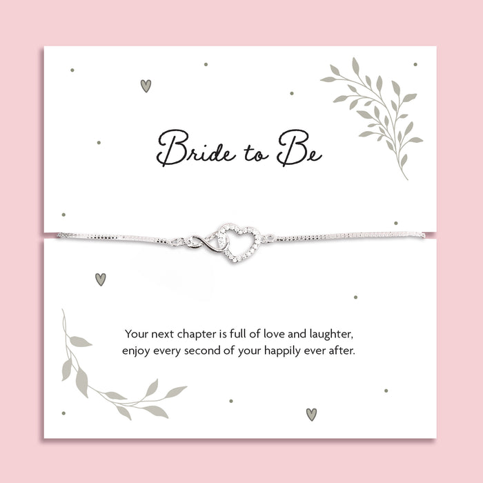 Silver zirconia heart and infinity charm bracelet displayed on a white card with the words "Bride to Be" and a romantic poem, symbolising love and celebration for an upcoming wedding. The bracelet is dainty and adjustable, styled on a soft pink background.