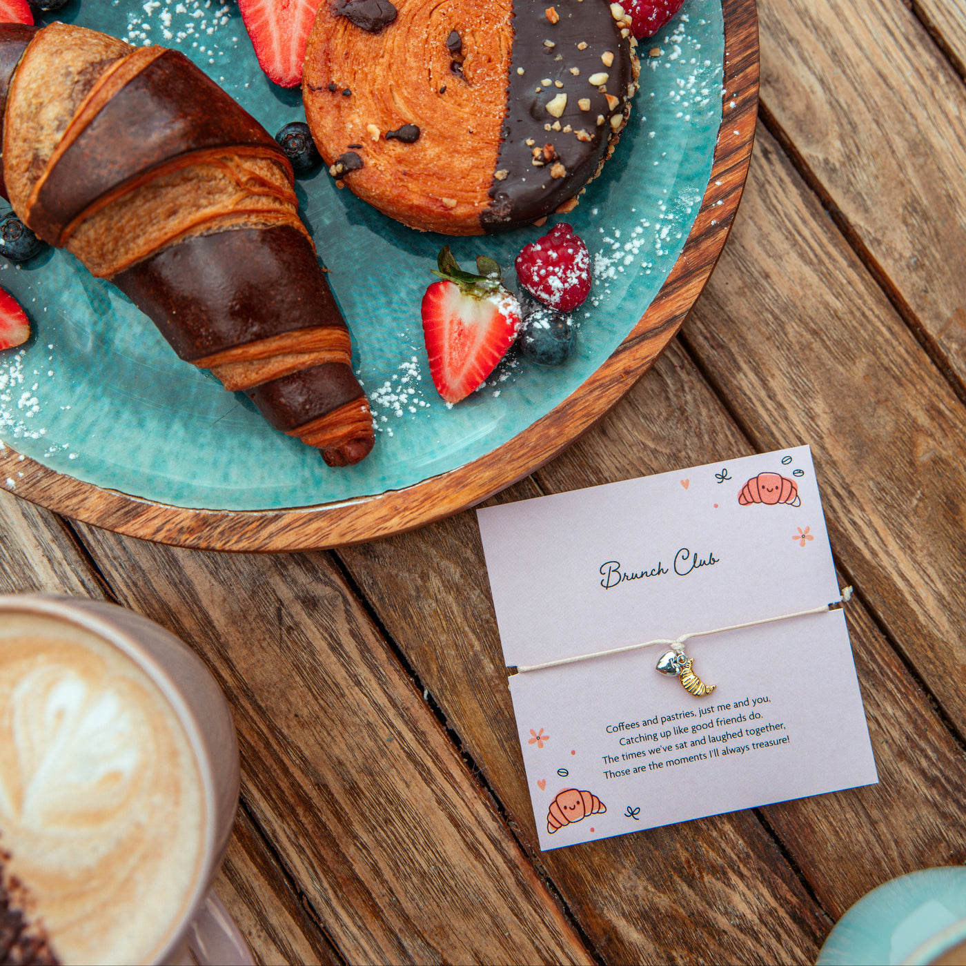 Dessert plate with pastries and berries on a wooden table, next to a coffee cup and a Brunch Club Bracelet and card.