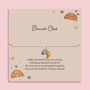 Brunch club friendship bracelet with gold croissant charm and silver heart on adjustable cord, displayed on brunch themed gift card
