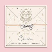 Adjustable beige cord bracelet with a silver 'Cancer' charm and small heart pendant, displayed on a zodiac-themed card featuring a crab illustration and the traits "Protective, Creative, Empathetic".