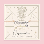 Adjustable peach cord bracelet with a silver 'Capricorn' charm and heart pendant, displayed on a zodiac-themed card featuring a goat illustration and traits reading "Reliable, Loyal, Patient".