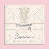 Adjustable peach cord bracelet with a silver 'Capricorn' charm and heart pendant, displayed on a zodiac-themed card featuring a goat illustration and traits reading 