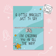 A silver bracelet with a heart and star charm displayed on a light blue greeting card that reads 'A LITTLE BRACELET JUST TO SAY' and 'I'M CHEERING YOU ON ALL THE WAY!'. The card is centered on a pink background with cute doodle illustrations.