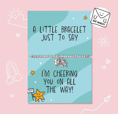 A silver bracelet with a heart and star charm displayed on a light blue greeting card that reads 'A LITTLE BRACELET JUST TO SAY' and 'I'M CHEERING YOU ON ALL THE WAY!'. The card is centered on a pink background with cute doodle illustrations.