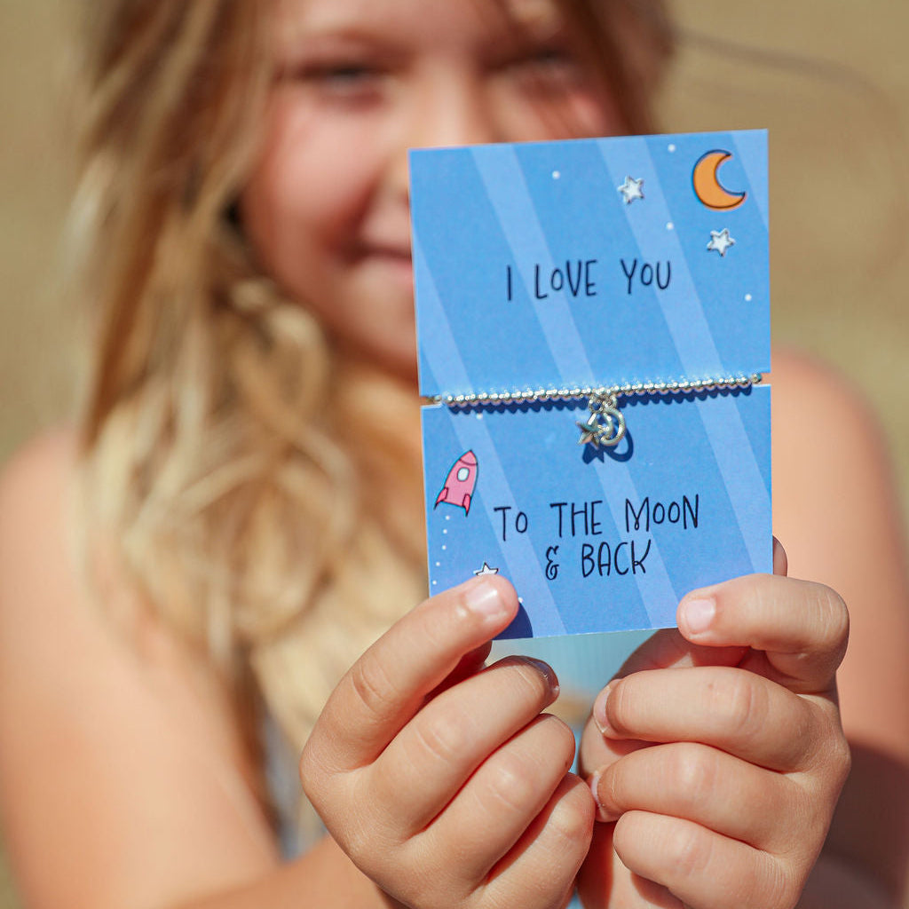 A child holds up a blue card featuring a beaded bracelet with a moon and astronaut charm. The card reads 'I LOVE YOU To THE MOON & BACK' and is photographed outdoors with the child blurred in the background.