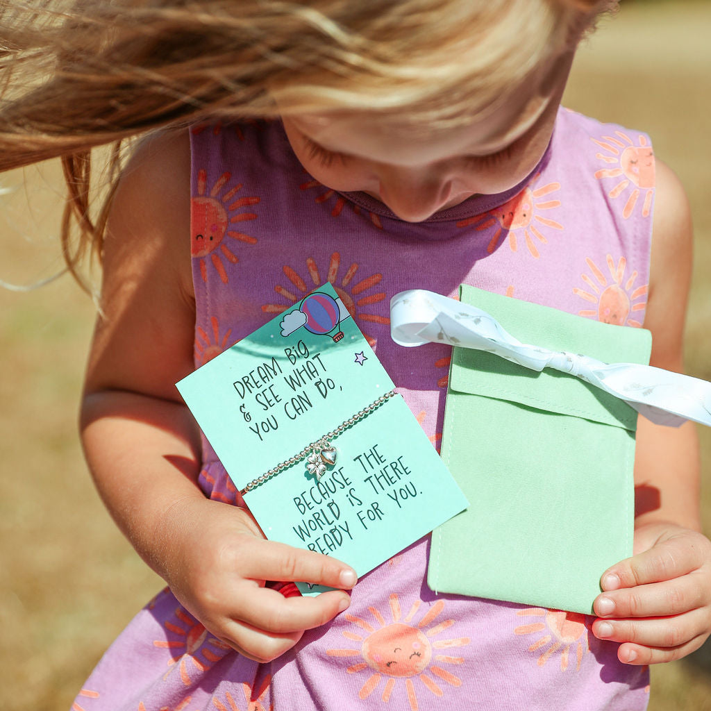 A young girl in a pink sun-print dress holds a mint green envelope and a light blue card with a good luck note and a small silver bracelet outdoors.