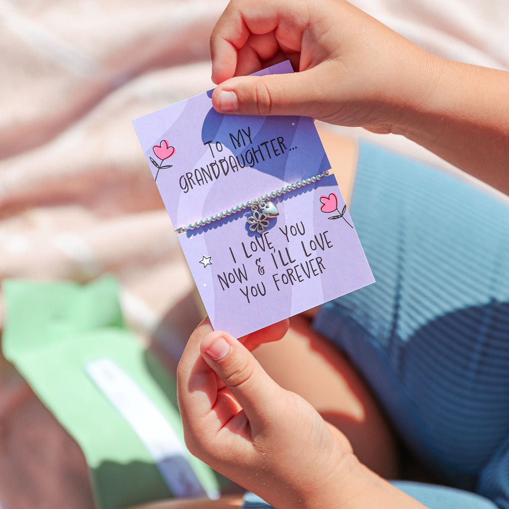 A child holding a small lavender card with a silver-toned bracelet attached, photographed outdoors on a sunlit blanket. The card reads, 'To my granddaughter.