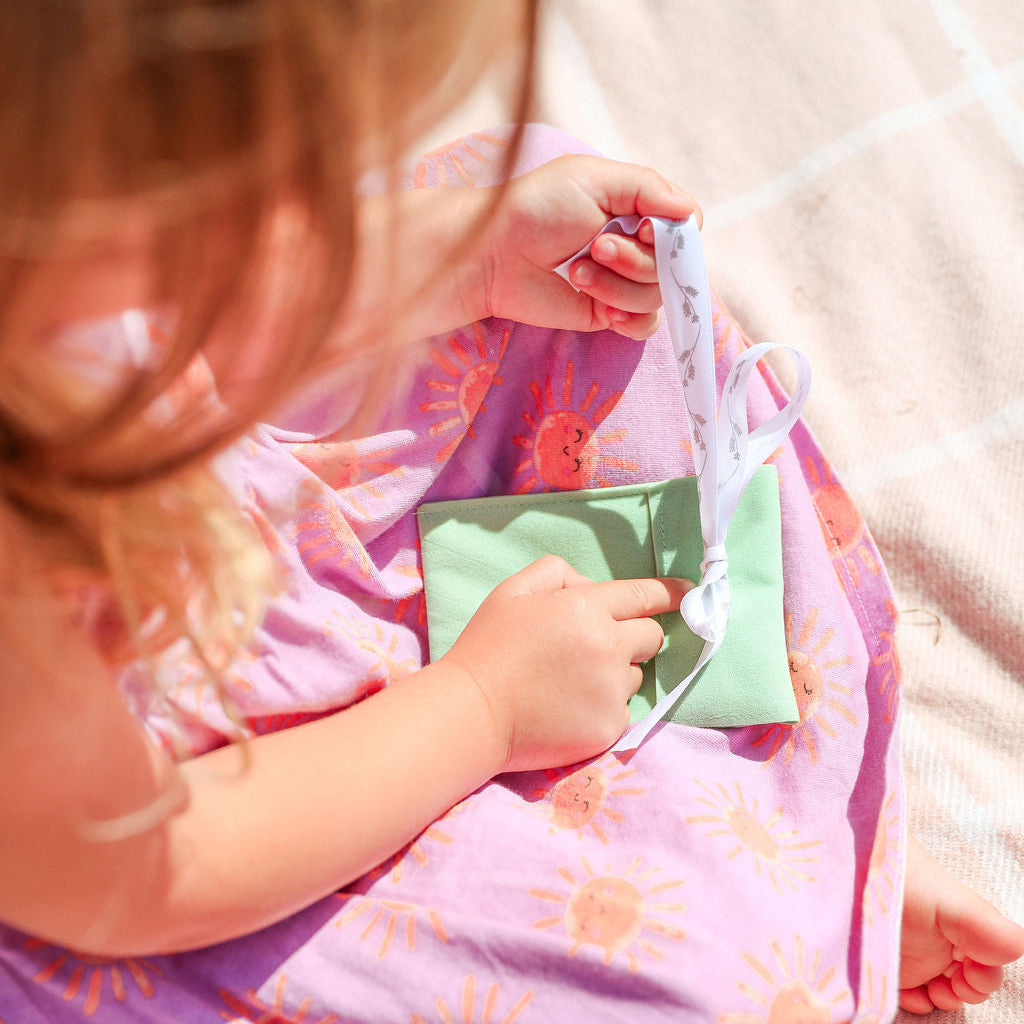 A child wearing a pink sun-patterned wrap or blanket sits outdoors on a sunlit beige fabric surface, holding a small mint green pouch with a white ribbon.