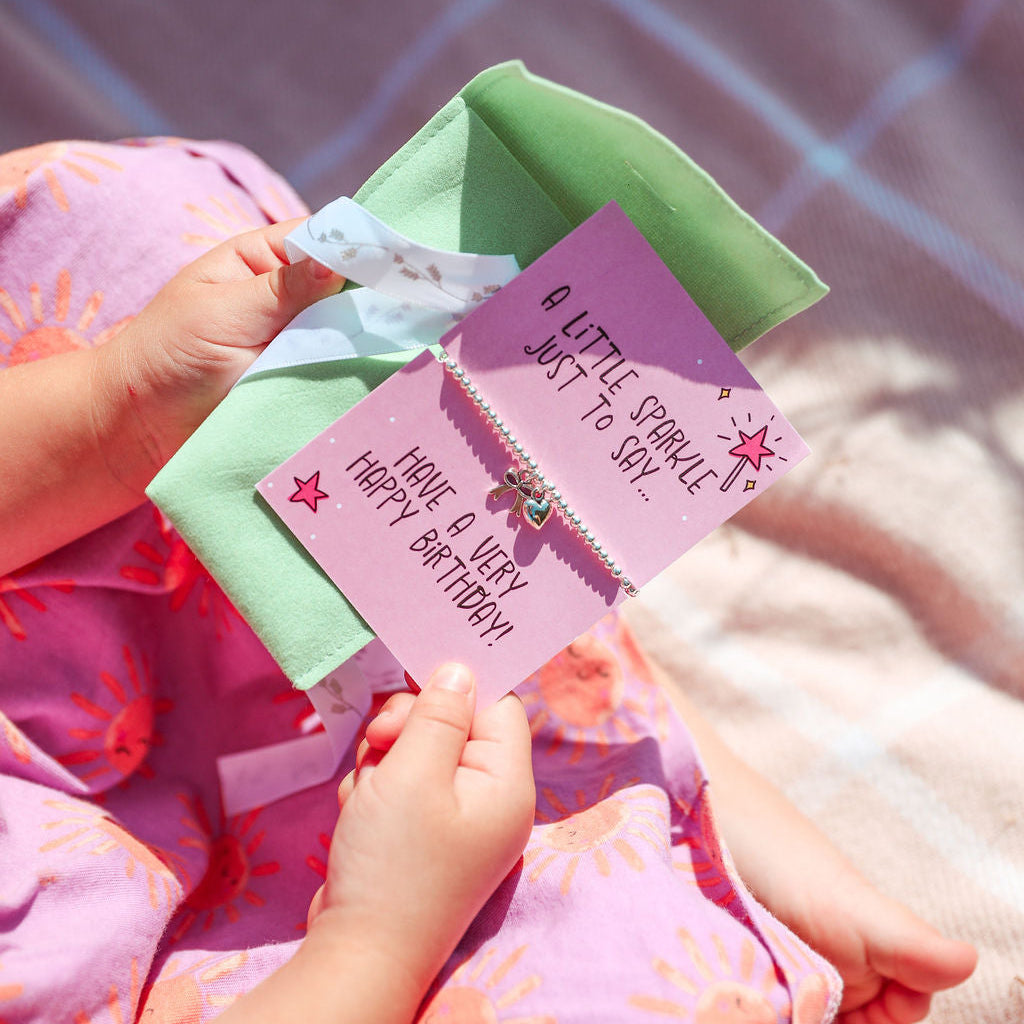 A child's hands hold a pink handwritten birthday motivational card with a beaded bracelet next to a green velvet envelope on a pink sun-print dress, sitting on a purple plaid blanket in bright sunlight.
