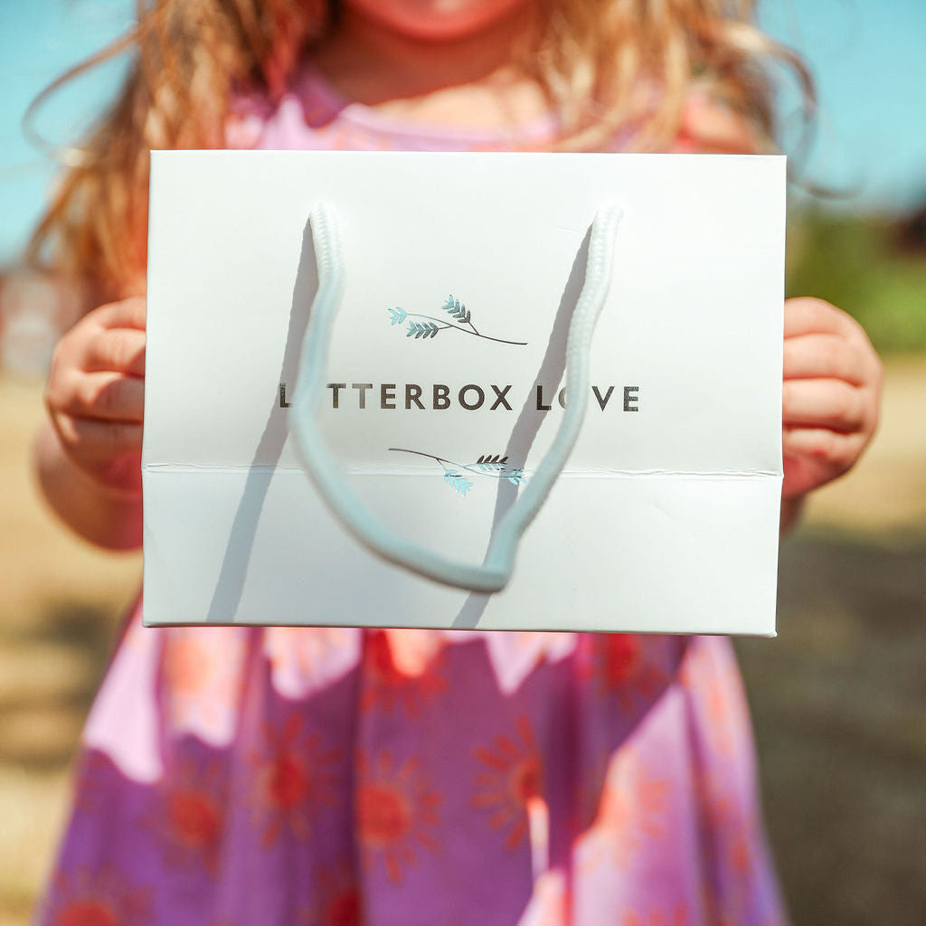 A young girl in a pink floral dress holds up a white Letterbox Love gift bag outdoors.