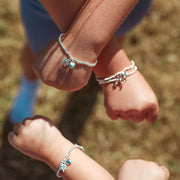 Three close-up views of kids' wrists and hands wearing delicate silver bracelets with small charms, including a heart and star, against a blurred outdoor background.