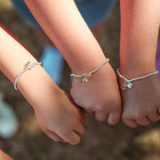 Three children's hands holding hands together, each wearing a delicate silver beaded bracelet with a small charm. The bracelets feature different charm designs, including a butterfly, a flower, and a small heart with a clear stone.