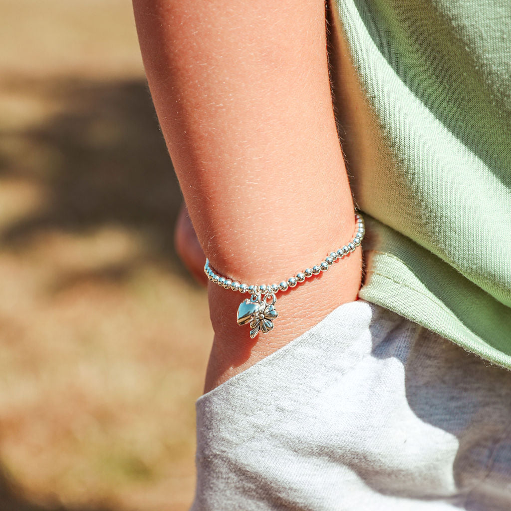 A close-up of a child's wrist wearing a silver beaded bracelet with a small blue and white butterfly charm, paired with a light green shirt and white shorts in an outdoor setting.