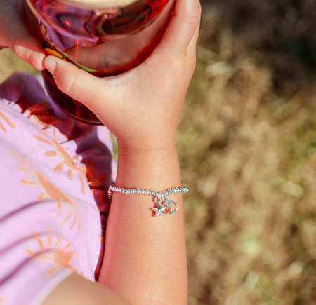 A close-up of a person's hand holding a glass jar with a bamboo lid, wearing a silver charm bracelet on the wrist. The bracelet features a small silver crescent moon and star charm.