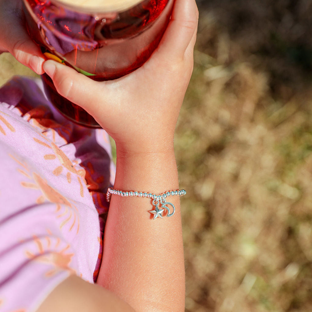 A close-up of a person's hand holding a glass jar with a bamboo lid, wearing a silver charm bracelet on the wrist. The bracelet features a small silver crescent moon and star charm.