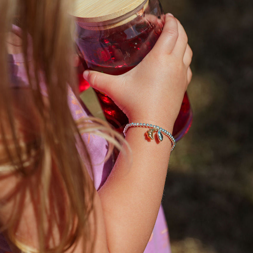 A close-up lifestyle shot of a child holding a glass jar outdoors, wearing a delicate silver beaded bracelet with a small gold charm on the wrist.
