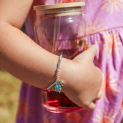 A close-up of a child's arm wearing a silver-toned beaded bracelet with a clover good luck charm, holding a glass jar with a wooden lid and red liquid, wearing a purple sun-patterned dress.