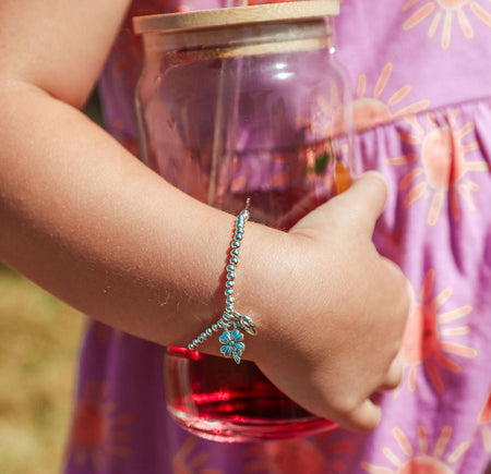 A close-up of a child's arm wearing a silver-toned beaded bracelet with a clover good luck charm, holding a glass jar with a wooden lid and red liquid, wearing a purple sun-patterned dress.